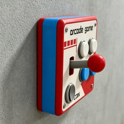 May include: A square light switch cover featuring a red and blue frame with a white faceplate. The design includes the text arcade game and coin, a red joystick, two white buttons, two blue buttons, and a gray button, all mounted on a wall.