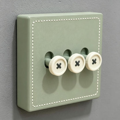 May include: A sage green light switch plate featuring a square shape with white stitched detailing along the border. The plate includes three protruding toggle switches designed to resemble round, cream-colored buttons with black cross-stitch patterns.